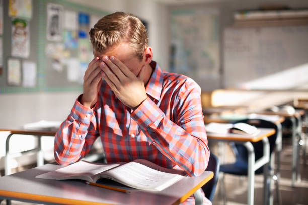 Stressed male student sitting with book at desk. Teenage boy is studying alone in classroom. He is at high school.